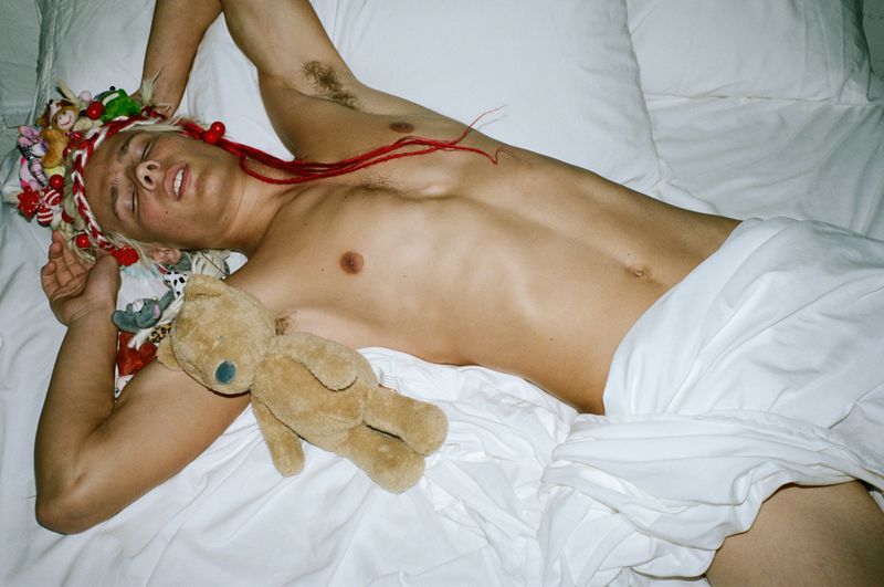Shirtless man lying in a white bed with a teddy bear and a floral crown for Parachute Mag