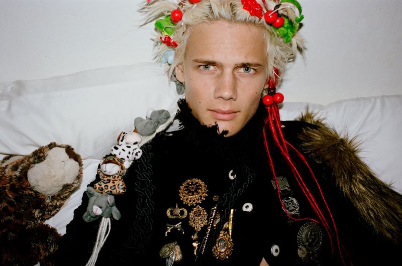 Model with blonde hair and cherry hair accessories wearing a black jacket adorned with pins against white pillows.