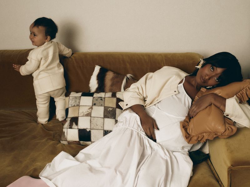 Pregnant woman in a white dress reclining on a gold sofa next to a standing child in cream clothing for Vogue Scandinavia.