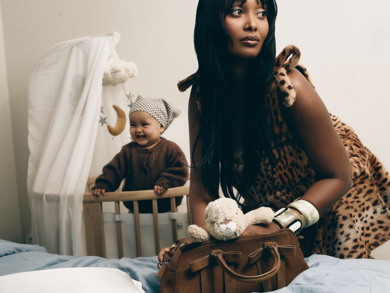 Woman in leopard print dress kneeling next to a smiling toddler in a crib inside a brightly lit bedroom.