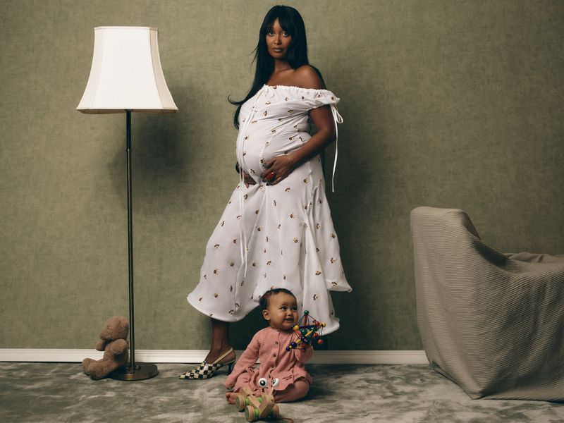 Pregnant woman in a white off-shoulder dress standing next to a baby playing on the floor with toys for Vogue Scandinavia.
