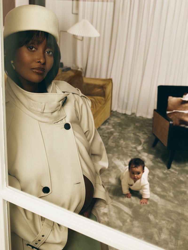 Pregnant woman in cream coat and pillbox hat with baby crawling on floor in living room by Vogue Scandinavia