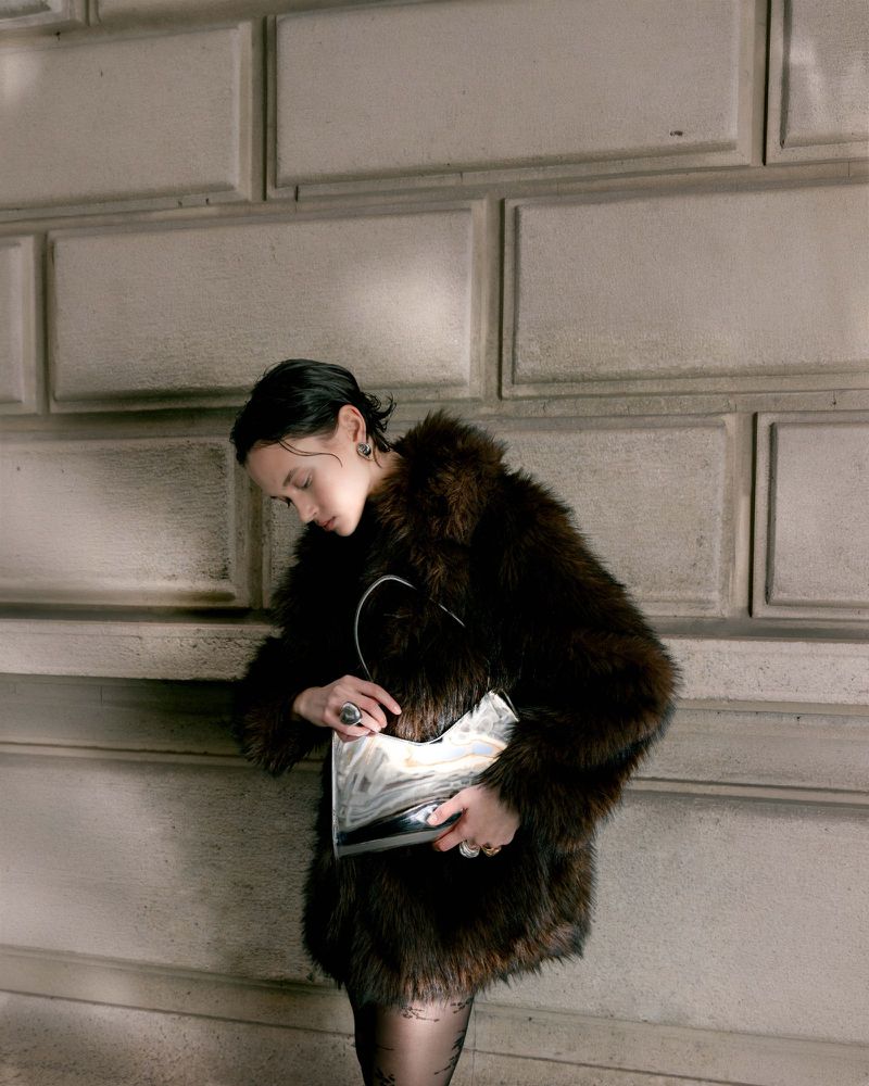 Model in brown faux fur coat holding a patterned handbag against stone blocks