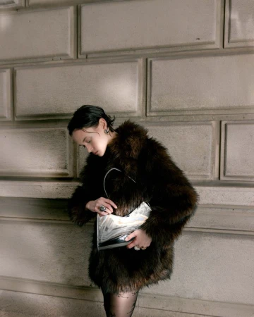 Model in brown faux fur coat holding a patterned handbag against stone blocks