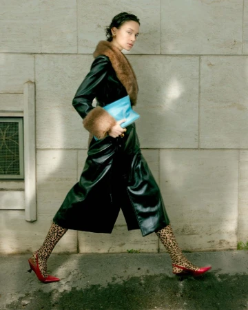 Model in a dark leather coat with fur trim walking along a tiled stone wall holding a bright blue clutch.