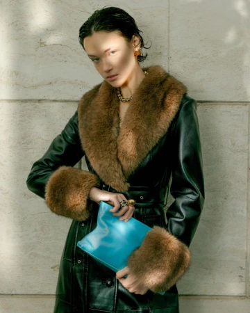 Model wearing a dark green coat with brown fur collar and cuffs holding a bright blue handbag against stone wall