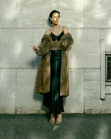 Model with gold face paint wearing a dark slip dress and faux fur coat against a stone wall