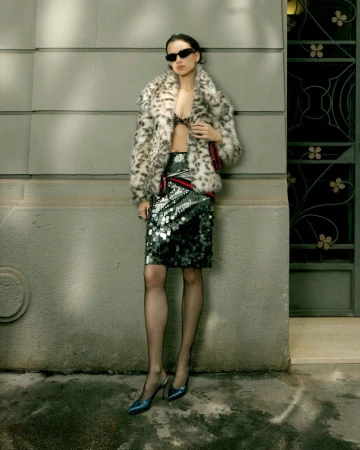 Model in sunglasses wearing a leopard print faux fur coat and sequin skirt leaning against an exterior wall.