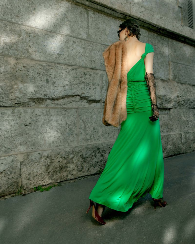 Model in vibrant green dress and fur stole walking past a stone wall for Harper's Bazaar Vietnam