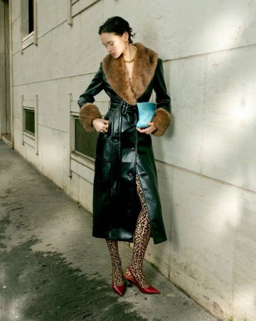 Model in a black leather coat with a brown fur collar standing against an urban stone building exterior.