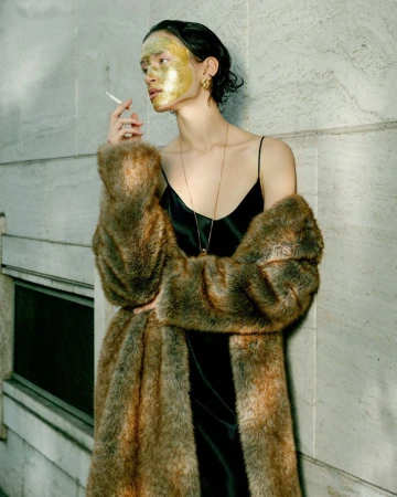 Model with gold face mask and cigarette wearing faux fur coat and black slip dress leaning against stone wall.