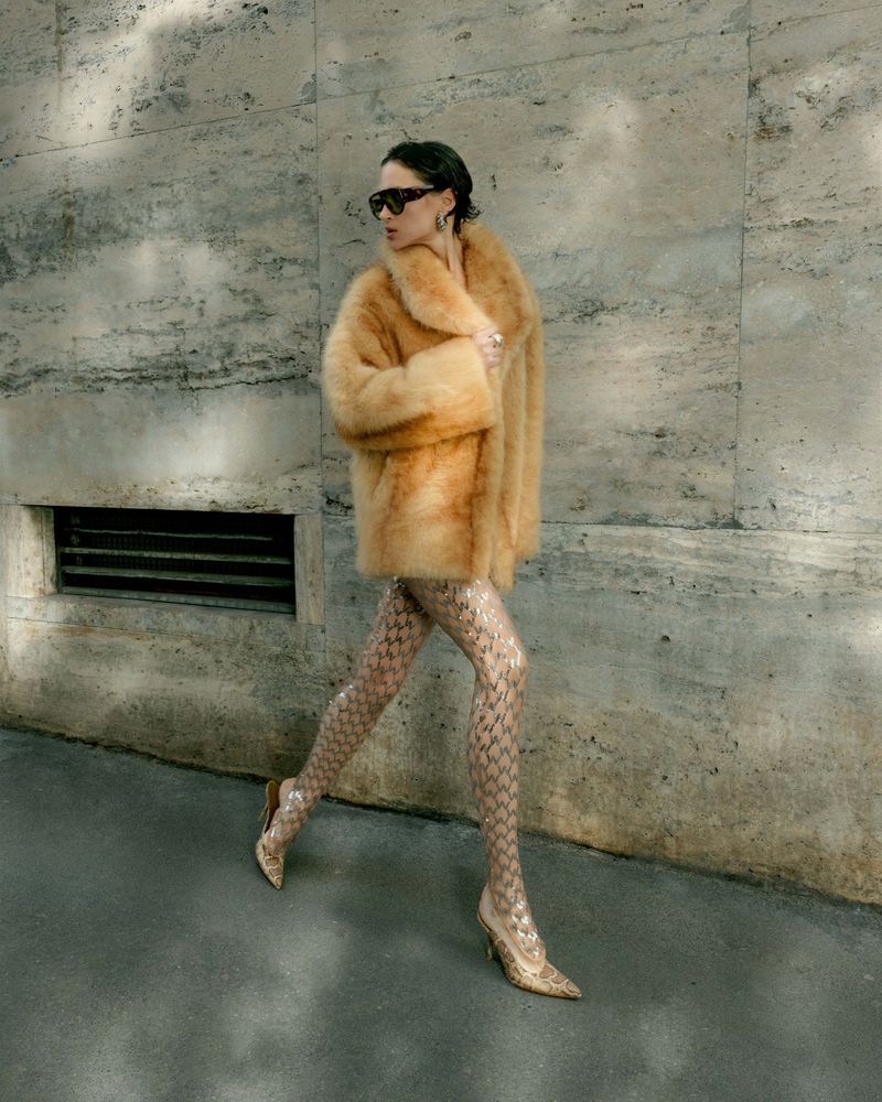Model in fluffy brown coat and patterned tights walking past a stone wall for Harper's Bazaar Vietnam