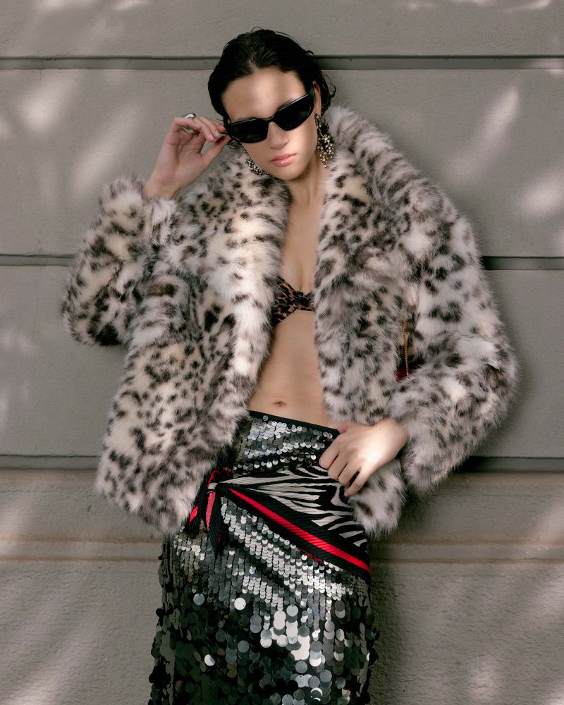 Model in leopard print coat and sequined skirt wearing sunglasses against a grey wall