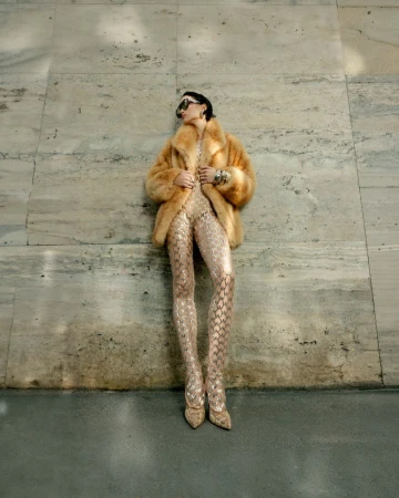Model in a faux fur coat and patterned jumpsuit leaning against a stone wall for Harper's Bazaar Vietnam city girl editorial.