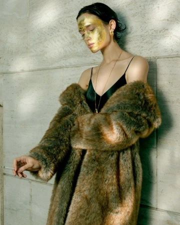 Model in faux fur coat and black slip dress with gold leaf makeup standing against a stone wall.
