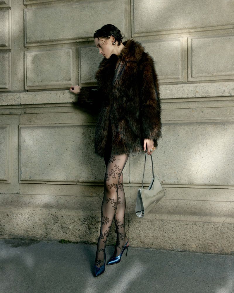 Model in dark faux fur coat and patterned tights leaning against a stone building exterior holding a metallic handbag