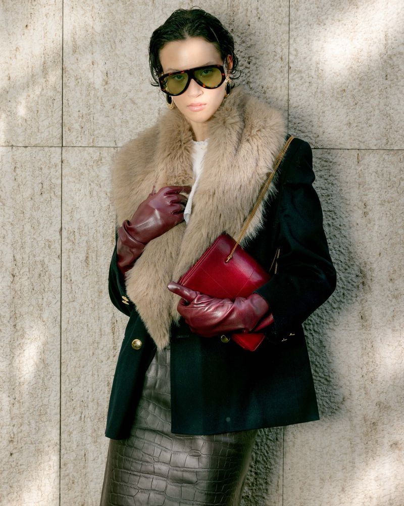 Model wearing dark sunglasses, fur collar coat, and burgundy leather gloves holding a red handbag against a stone wall.