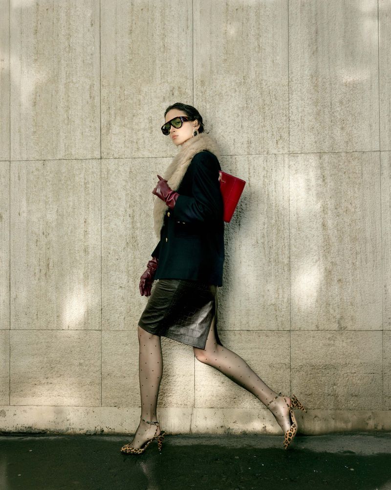 A stylish woman strides confidently in a sleek black blazer, leather skirt, and leopard heels, carrying a bright red clutch against a concrete wall.