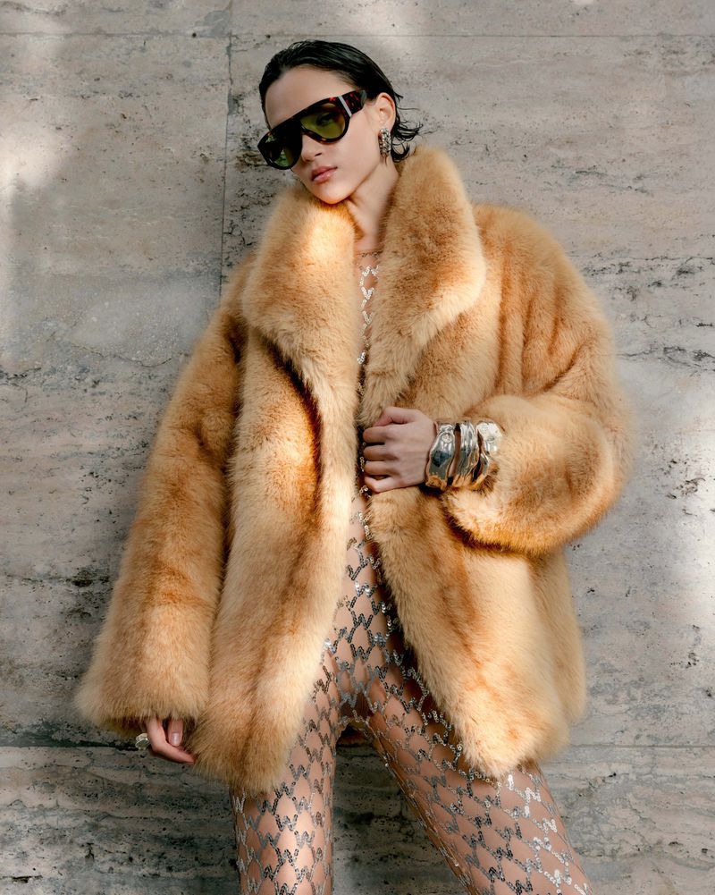 Model in oversized faux fur coat wearing sunglasses and patterned tights against a textured stone wall.