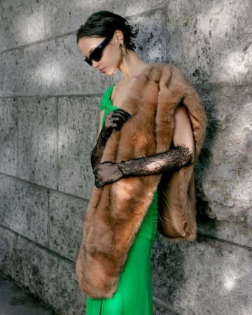 Model in green dress and fur stole wearing sunglasses leaning against a stone wall