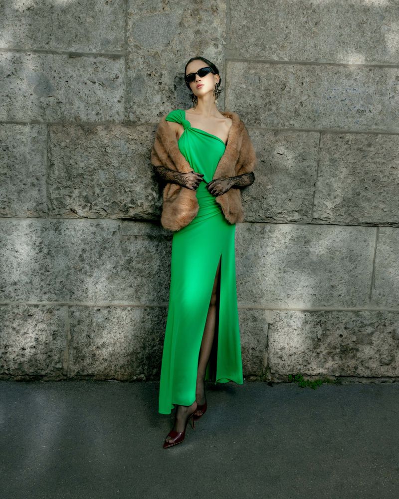 Model wearing a bright green dress, fur stole, and sunglasses leaning against a rustic stone wall