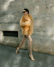 Model in fluffy brown coat and patterned tights walking past a stone wall for Harper's Bazaar Vietnam