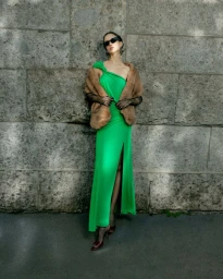 Model wearing a bright green dress, fur stole, and sunglasses leaning against a rustic stone wall
