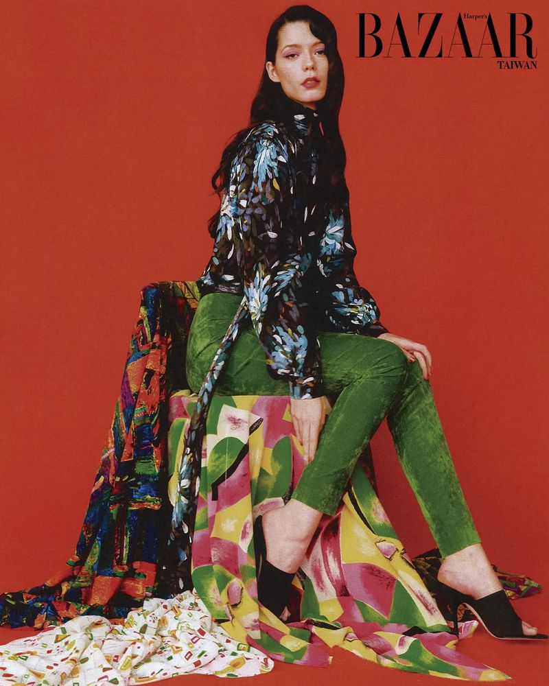 Model seated on a patterned chair wearing a floral blouse and green trousers against a solid red studio background.