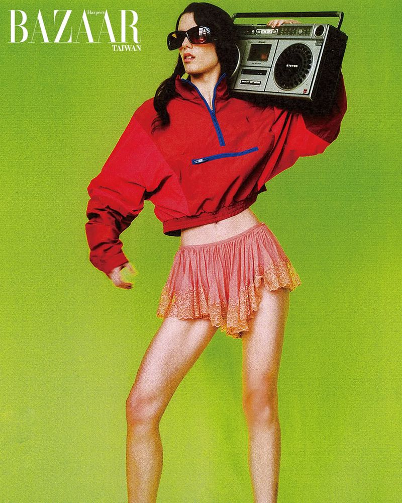 Model wearing a red half-zip jacket and pink ruffled shorts holding a vintage boombox against a bright green background.