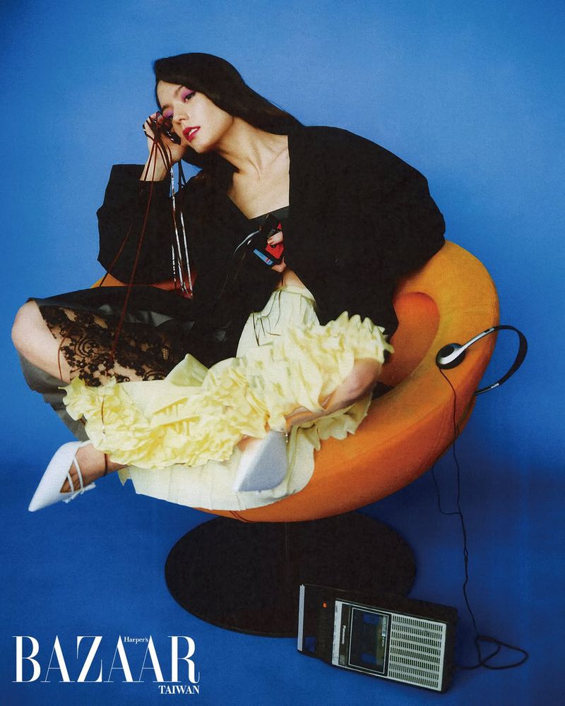 Woman in black jacket and ruffled yellow skirt seated in an orange bowl chair against a blue background for Harper's Bazaar Taiwan.