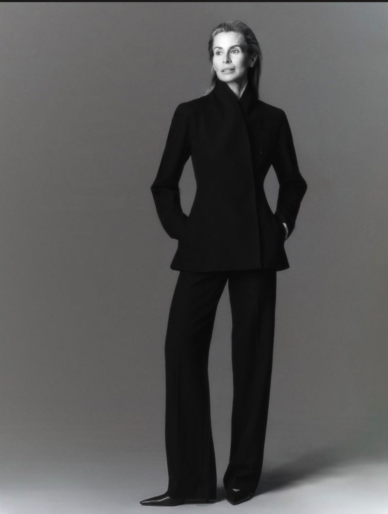 Model Niki Taylor standing in a tailored black jacket and matching trousers against a neutral grey studio backdrop.