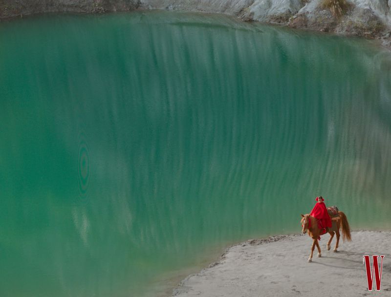 A person wearing a bright red cape riding a brown horse along a sandy shore next to a large body of turquoise water.