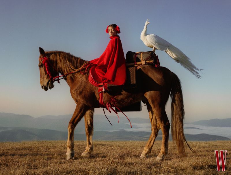 Young model in vibrant red cape riding a brown horse with a white peacock perched behind in a mountain landscape.