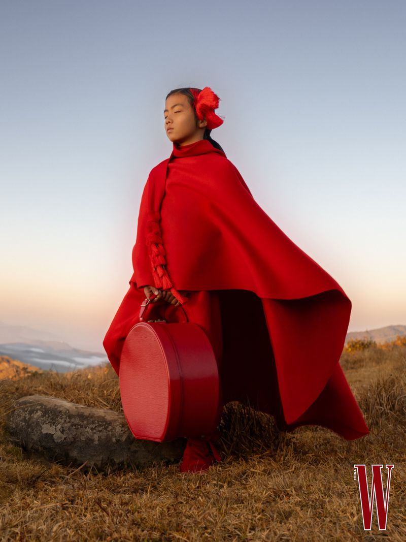 Model in a vibrant red cape holding a matching red hat box outdoors against a mountain landscape at sunset.