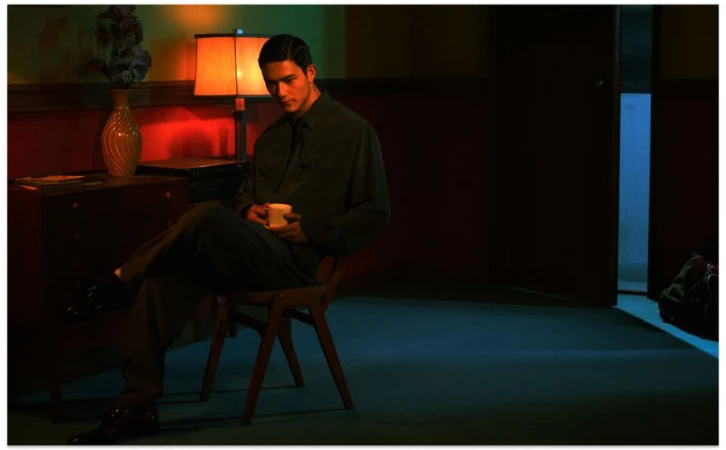 Man seated on a wooden chair holding a small cup in a dimly lit room with warm and cool lighting by Leslie Zhang for Vogue China Men.