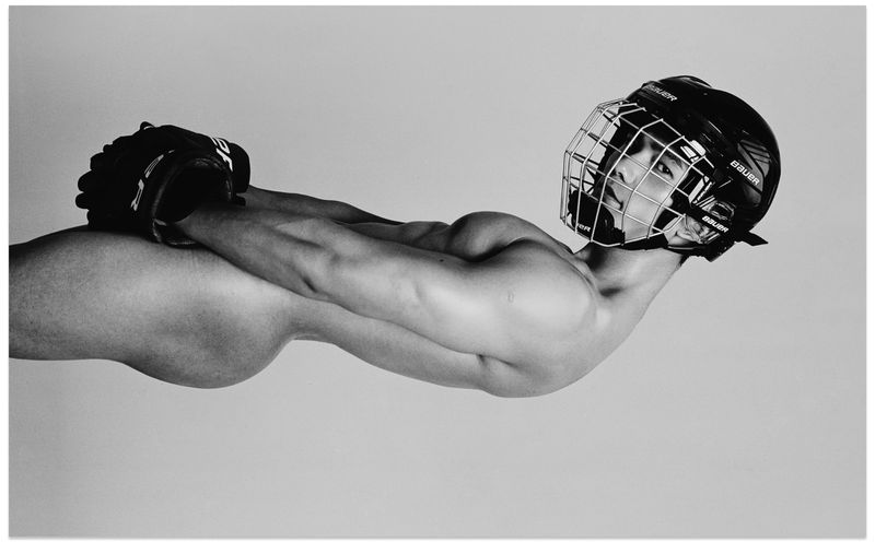 Monochrome studio portrait of a shirtless male athlete wearing a hockey helmet and holding hockey gloves.