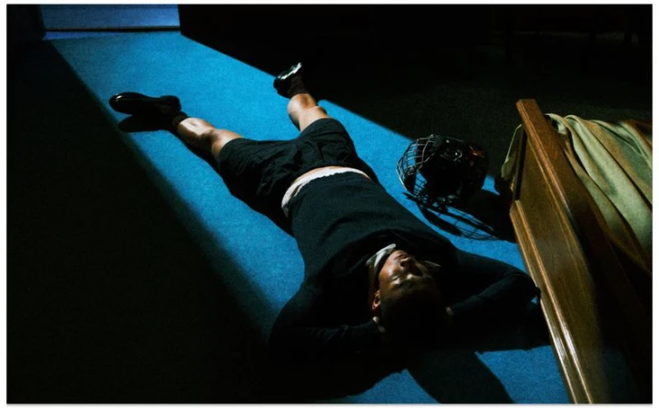 Man lying face down on a blue carpeted floor next to a sports helmet in a moody interior illuminated by a beam of light.