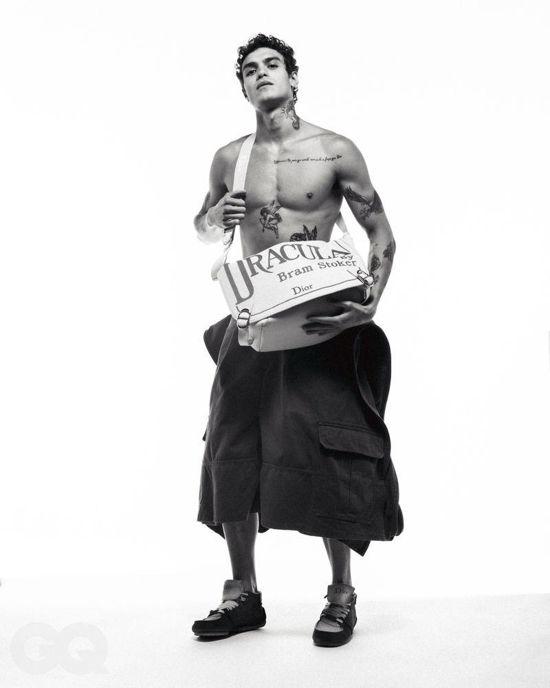 Shirtless male model wearing dark cargo shorts and holding a white crossbody bag against a plain white background