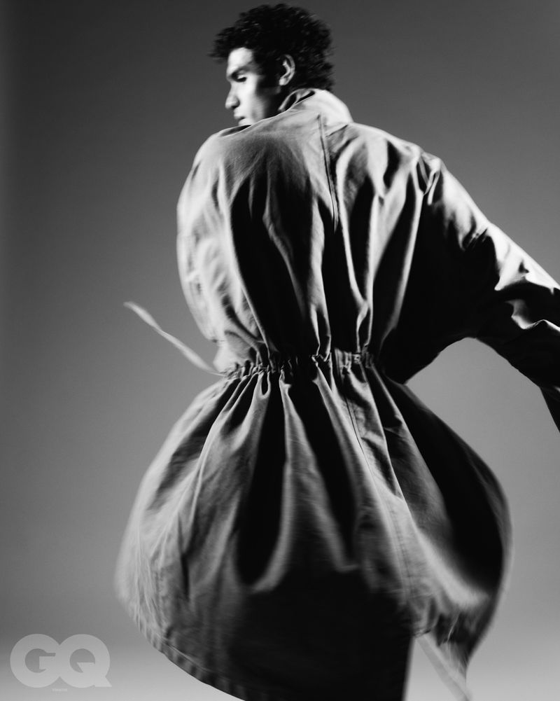 Monochrome fashion editorial image of a male model in motion wearing a flared coat against a plain gradient background.