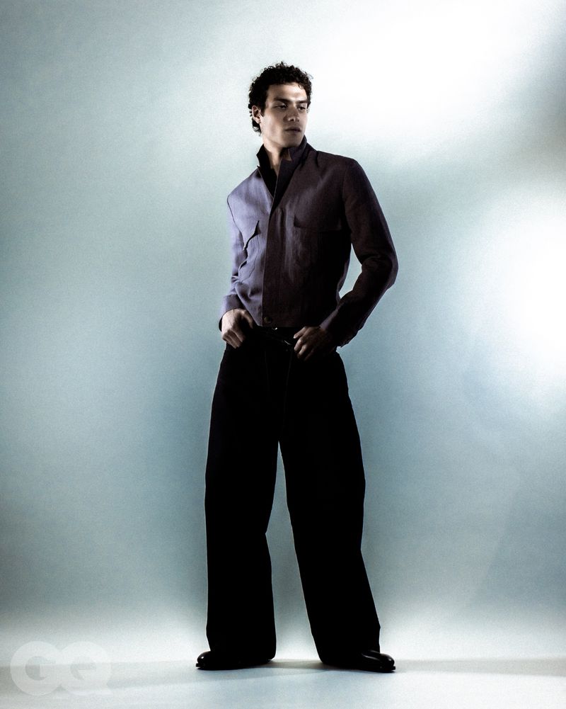 Male fashion model poses indoors wearing a purple button-down shirt and large black trousers in a studio setting with even lighting.