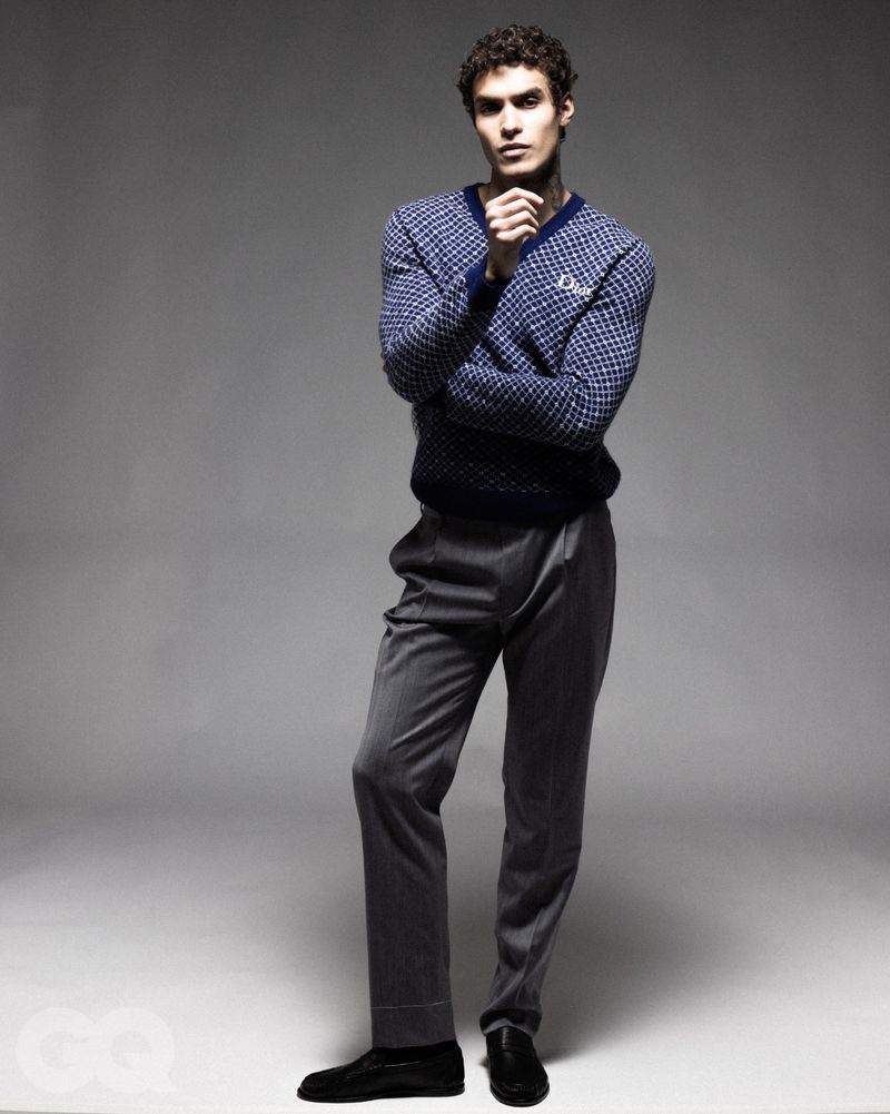 Full-length portrait of a male model wearing a patterned blue v-neck sweater and grey trousers against a seamless grey studio background.