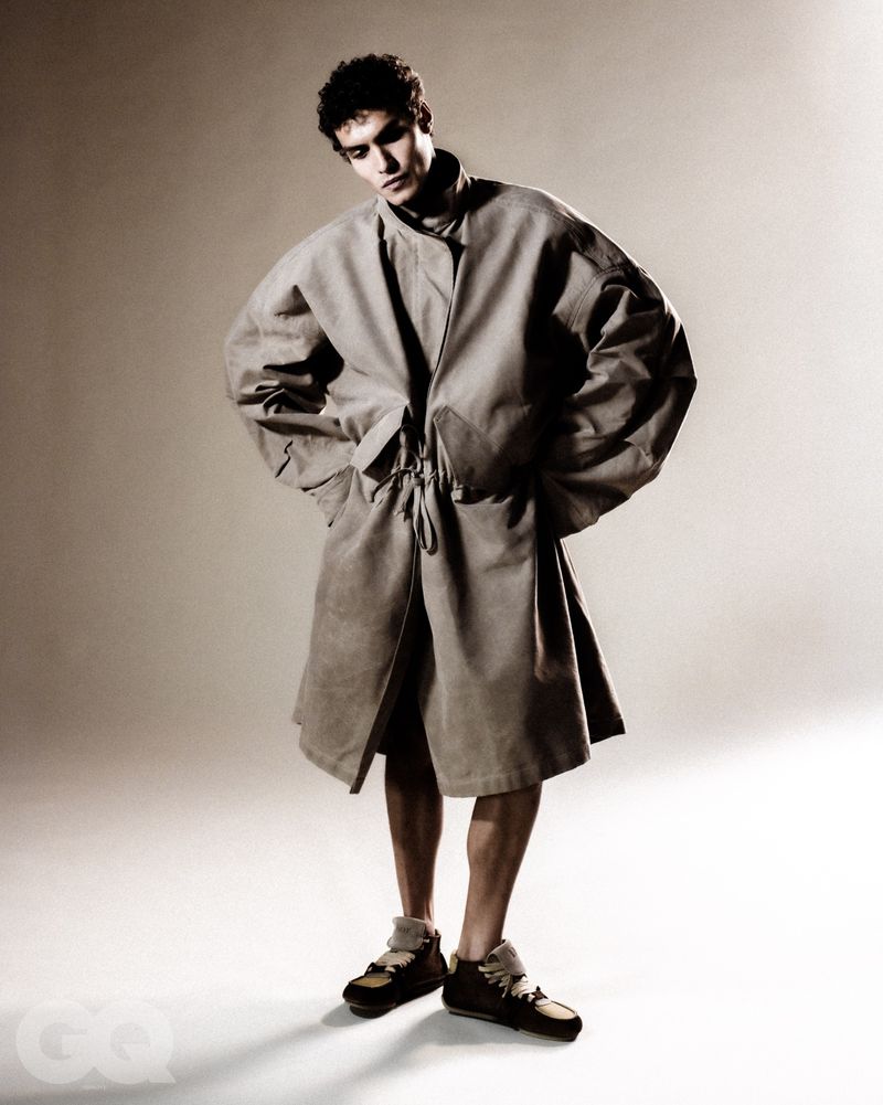 Male model wearing a voluminous oversized coat and dark sneakers standing against a plain gradient studio background