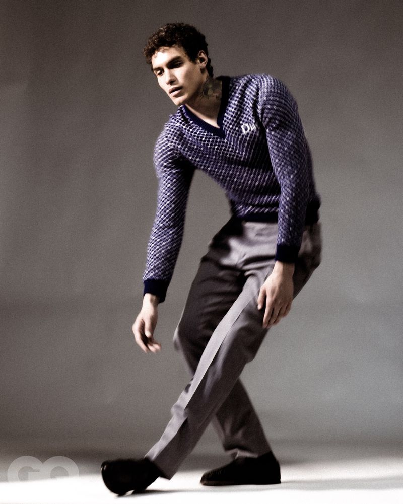 Male model wearing a patterned blue V-neck sweater and grey trousers posing against a grey studio backdrop.