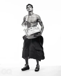 Shirtless male model wearing dark cargo shorts and holding a white crossbody bag against a plain white background
