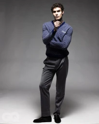 Full-length portrait of a male model wearing a patterned blue v-neck sweater and grey trousers against a seamless grey studio background.