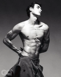Monochrome portrait of a shirtless male model with tattoos posing against a grey backdrop for GQ Turkey