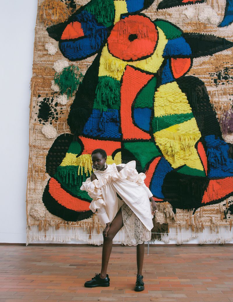 Model Yar Aguer standing in a ruffled white top and lace skirt before a large colorful textured wall hanging