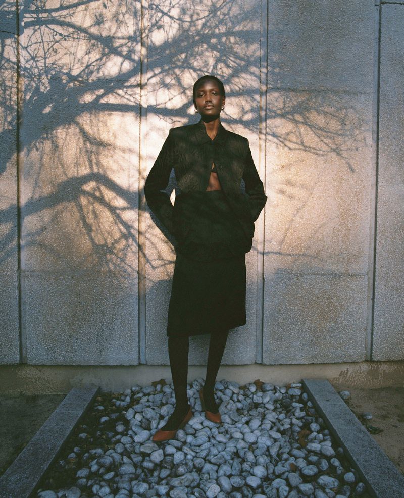 Model standing against a textured stone wall with cast tree branch shadows for a Vogue Espana fashion editorial