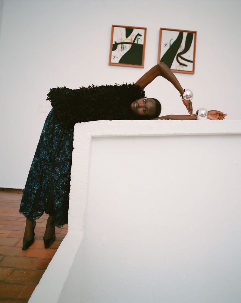 Model Yar Aguer in a textured black top and dark skirt leaning on a white partition with two framed abstract paintings on the wall behind.