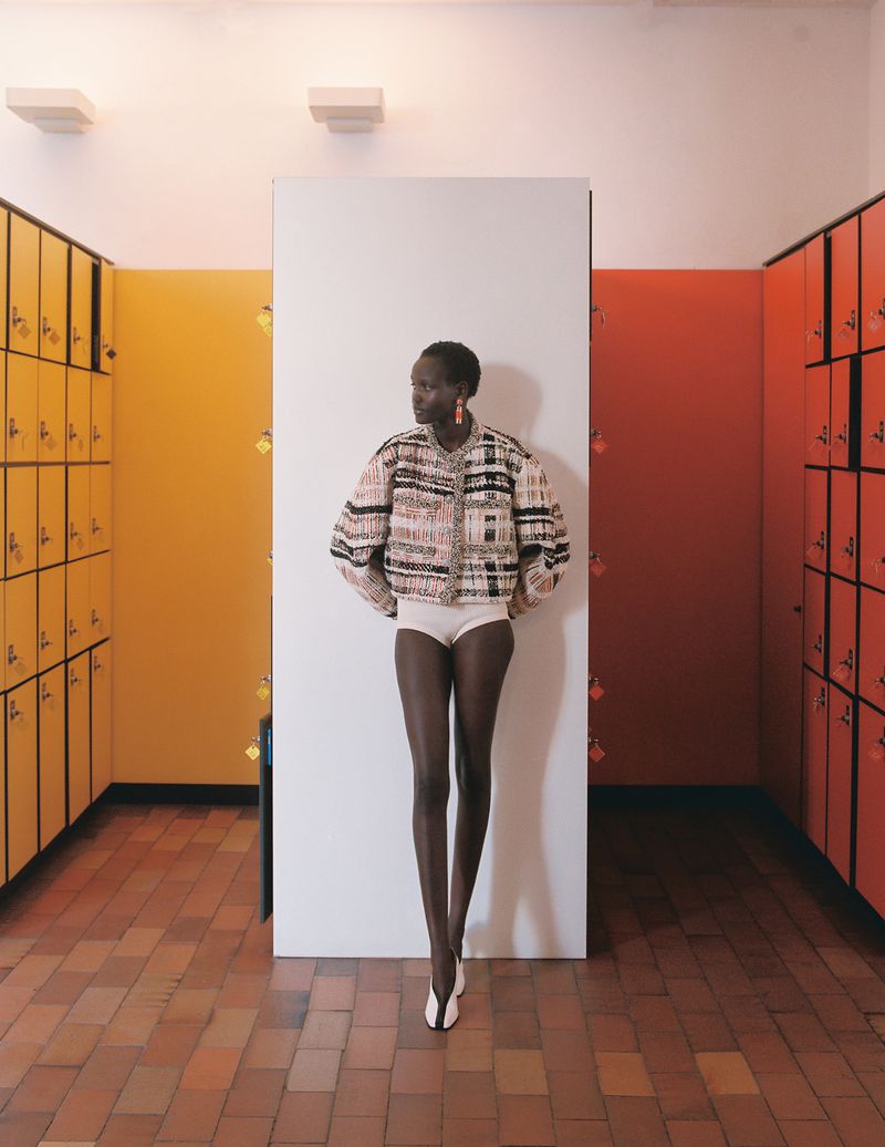 Fashion shot of a Black woman wearing a patterned jacket and briefs, standing in front of a white panel, in a locker room setting with orange and yellow lockers.